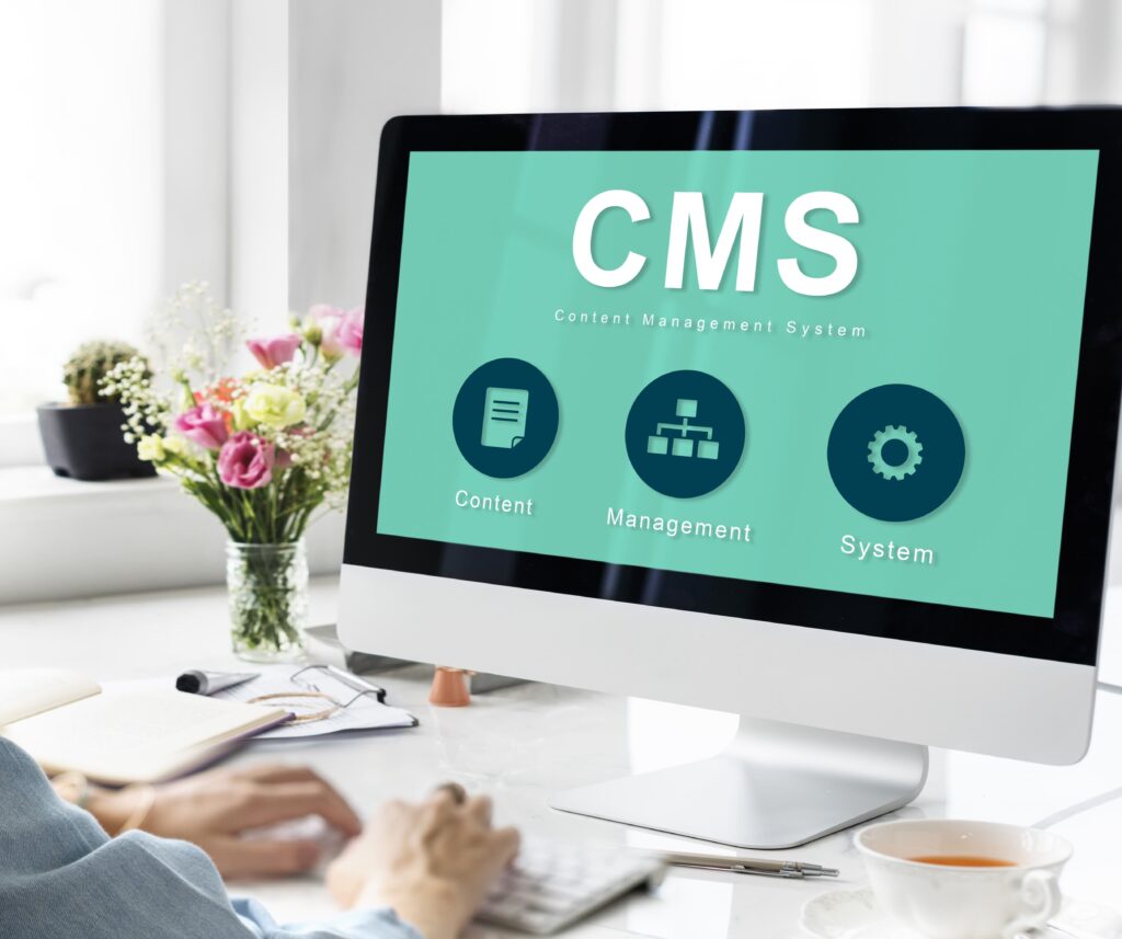 Монитор с интерфейсом системы управления контентом (CMS) на экране. Пользователь работает за компьютером, на переднем плане — цветы, блокнот и чашка чая.